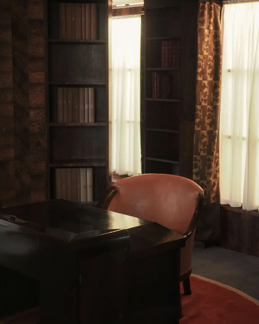 Mesa de madeira escura, poltrona de couro e estantes de livros, com a luz da janela ao fundo.